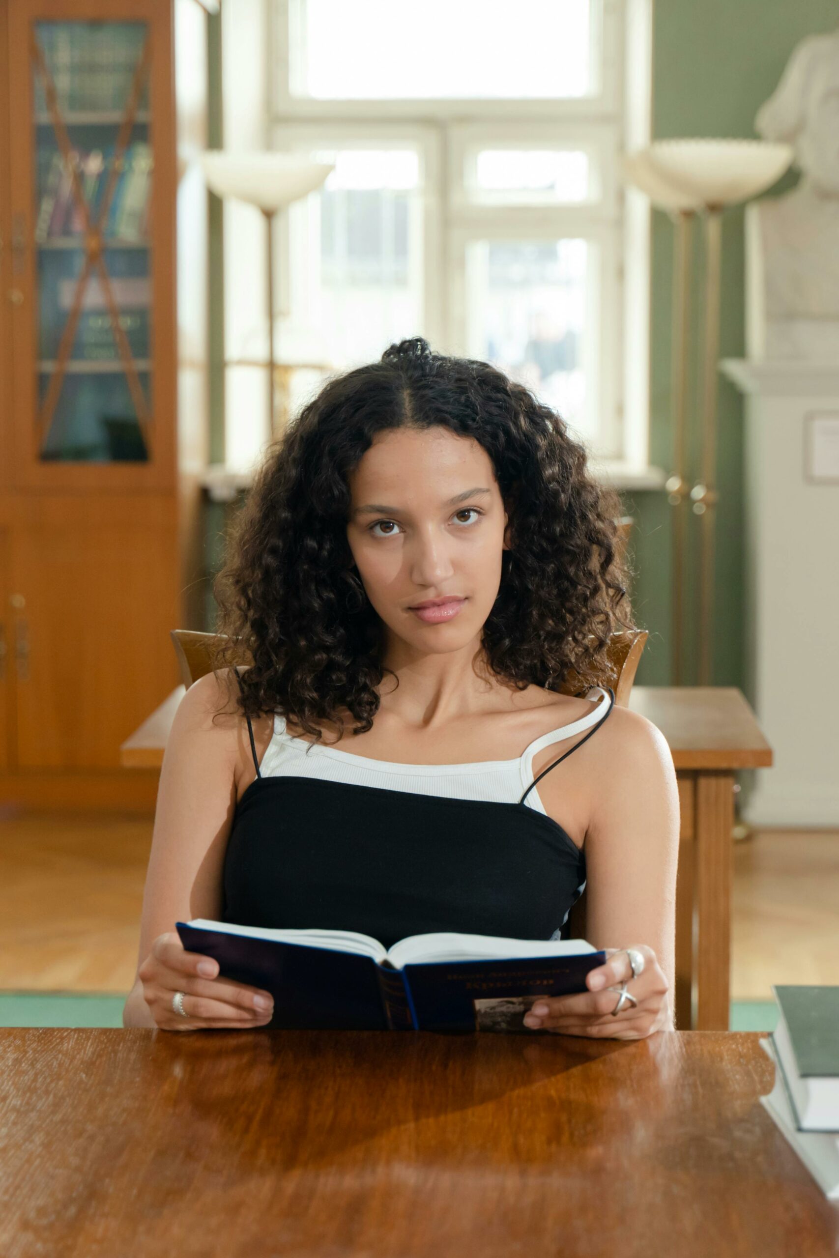 A woman with curly brown hair is sitting at a table in a library. She is looking up at the camera but not smiling, and is wearing black, thin-strapped top over a white thin-strapped top. She has an open book in her hands.