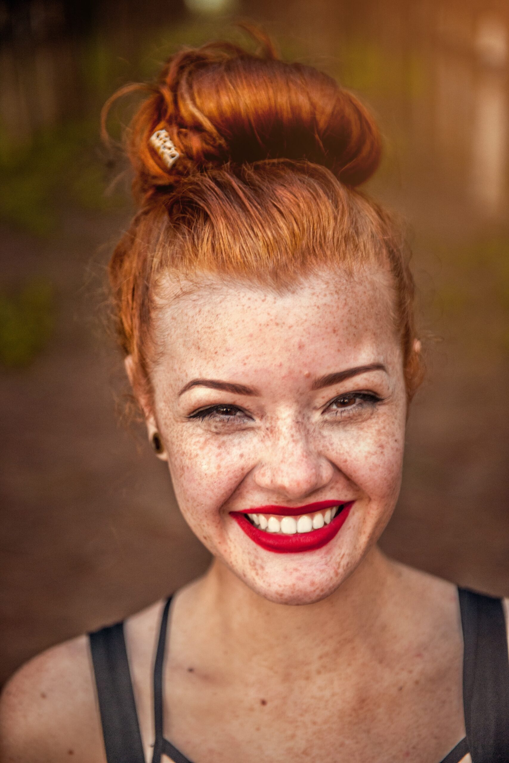 A close-up photograph of a young girl with vibrant red hair piled casually on top of her head. She has a scattering of freckles across her face, neck and shoulders. She wears bright red lipstick, which accentuates her wide smile as she looks directly at the camera with a cheerful expression.